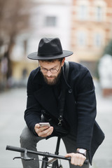 Carefree time in the city. Low angle view of serious young bearded man holding and typing mobile phone while sitting outdoors