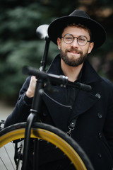 Bearded man with a fixie bicycle. Low angle view of confident young bearded man carrying his bicycle on shoulder and looking at camera while walking outdoors.