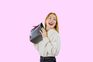 A portrait of a beautiful cute red-haired girl who holds a gift bag in a black box with a red velvet ribbon raising a gift, demonstrates his smile, looking enthusiastically to the side against an