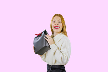 Portrait of a beautiful cute red-haired girl holding a gift bag in a black box with a red velvet ribbon raising a gift, demonstrating his smile, looking enthusiastically at the camera on an isolated