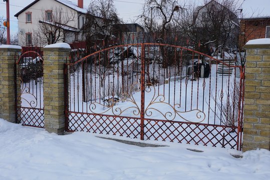 red metal gates of forged rods with a pattern and part of a brick fence on the street in white snow