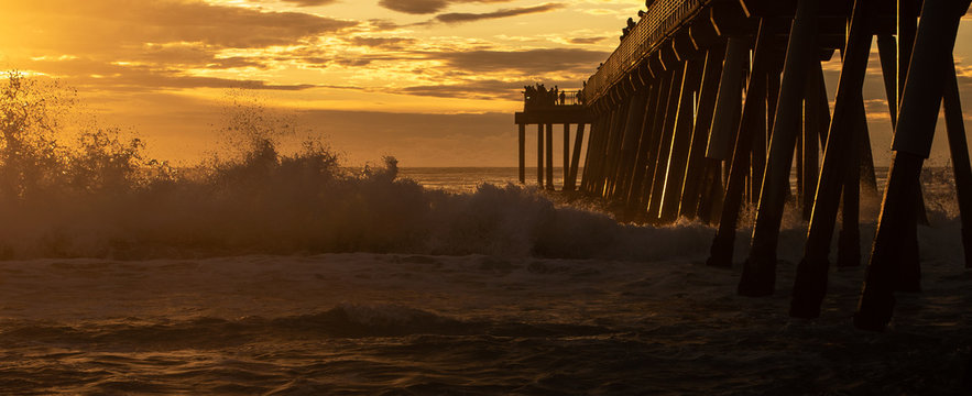 Fototapeta Waves breaking under the pier at Hermosa Beach, California. Tourists and visitors silhouetted against cloudy sunset sky