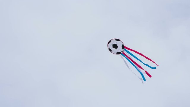 Kite in the Form of a Soccer Ball. An air kite painted under a soccer ball with multi-colored ribbons soars in the sky. Filmed at a speed of 240fps