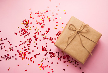 Gift box wrapped in brown colored craft paper and tied with rope on pink background with heart shape red confetti.