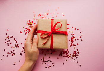 Gift box wrapped in brown colored craft paper and tied with red bow on pink background with red confetti. Lady's hand holding a giftbox.