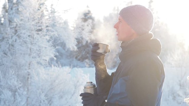 Hiking winter forest concept. Young man drinking tea or coffee in snowy winter forest. Mountain camping concept. Traveler man hiking in forest in winter time using thermos holding cup of tea.