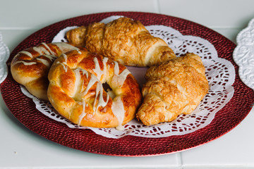 Fresh croissants and buns on round plate on white wooden table.