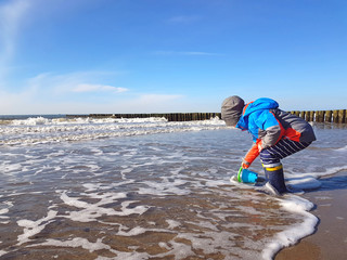 Boy child playing in the waves on Baltic Sea beach during winter vacation in german coast. Kid play in ocean sand dunes on cold autumn or spring day.