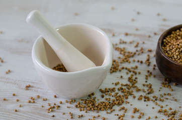Coriander seeds in porcelain mortar and on white wooden table.