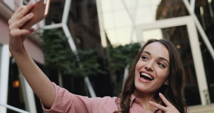 Close up of the pretty young brunette girl taking selfie photos on the smartphone device camera and posing to it. Outdoor.