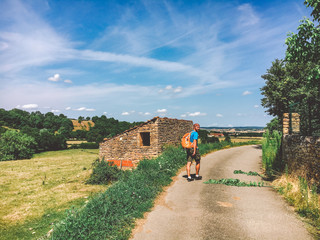 A man a hiker walks along an asphalt rural road outside the city in France, a region of Burgundy, which is bathed with an orange backpack in the summer hot sunny weather - Powered by Adobe