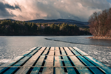 Fototapeta premium Mole (pier) on the lake. Wooden bridge in forest in winter time with blue frozen lake. Lake for fishing with pier. Dark lake (ice) with hills
