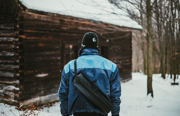 Photo of adventurer (hiker)  in snow covered forest in front of abandoned cottage (view from his back). Hiker Man in blue jacket standing in mountains with old destroyed cottage on background.