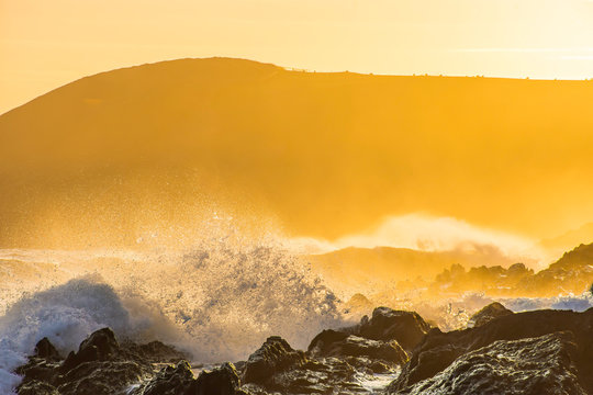 Sunset on Pembrokeshire coast.Big waves splashing on rocky shore and mist over sea with cliff in background during golden hour.