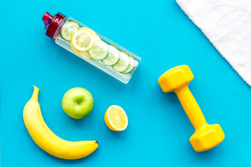 Healthy fruit water for sport, fitness. Bottle of water with lemon and cucumber near sport equipment dumbbells on blue background top view