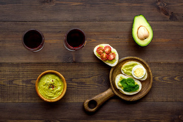 Light appetizer with avocado. Toasts with vegetables and guacamole near glasses of wine on dark wooden background top view