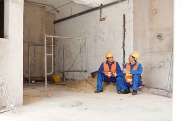 Two positive construction wokers in hard hats drinking coffee and chatting during short break