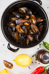 Spicy mussels in black saucepan. White wooden background decorated with lemon, hot peppers and greens