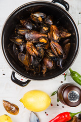 Spicy mussels in black saucepan. White wooden background decorated with lemon, hot peppers and greens