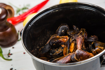 Spicy mussels in black saucepan. White wooden background decorated with lemon, hot peppers and greens