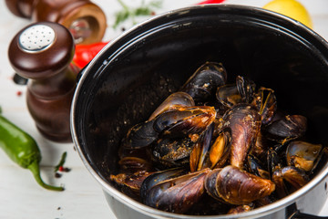 Spicy mussels in black saucepan. White wooden background decorated with lemon, hot peppers and greens