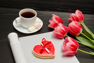 Holiday concept. Bouquet of pink tulips, a cup of coffee, red heart-shaped cookies with a note, empty magazine on a black wooden background