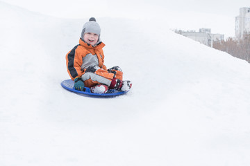 Fototapeta na wymiar Happy laughing small boy slides down the hill on snow saucer. Seasonal concept. Winter day.
