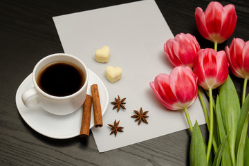 Fototapeta na wymiar Holiday concept. Bouquet of pink tulips, a cup of coffee, heart-shaped sugar, cinnamon, star anise and sheet of paper on a black wooden background. Close-up.