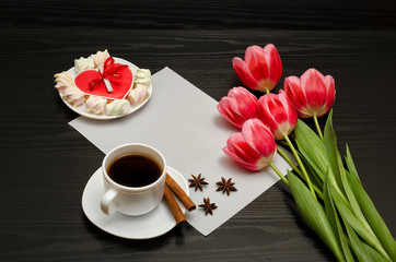 Fototapeta na wymiar Holiday concept. Bouquet of pink tulips, a cup of coffee, red heart-shaped cookies with a note, cinnamon, star anise and sheet of paper on a black wooden background