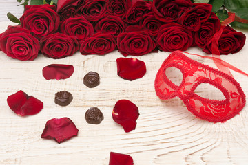 Romantic carnival concept. Red carnival mask, bouquet of red roses and chocolates on light wooden background.