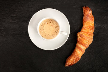 A photo of a croissant with a cup of coffee, shot from above on a black background with copy space