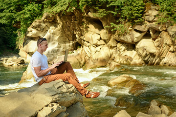 Work in nature concept. Man with notebook sitting at the bank of river over the waterfall and green trees. Side view