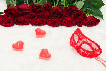 Romantic carnival concept. A red carnival mask and a bouquet of red roses and a heart-shaped candle on white fur.