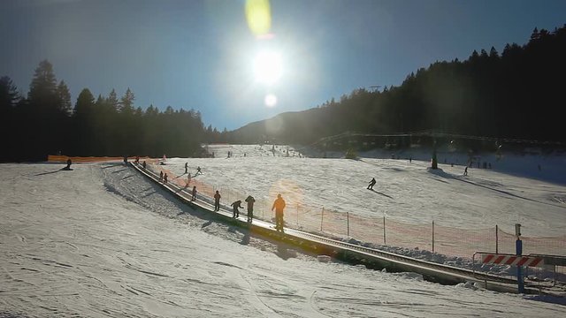 Skiers reaching the top of a descent on a ski slope with a tapis roulant (automated carrying track). Zooming-out shot, daylight, with a warm sun and flares.