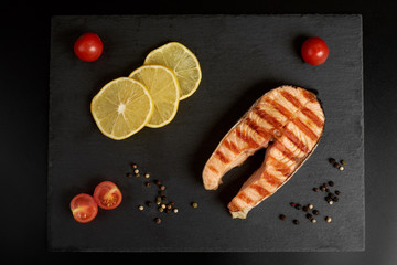 Grilled salmon steak with a pepper, lemon and tomatoes on a slate plate.