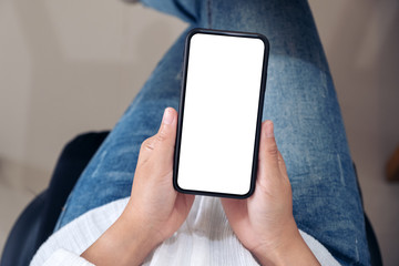 Top view mockup image of hands holding black mobile phone with blank white screen while sitting