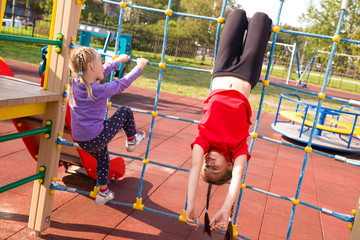 Two happy caucasian girls having fun on playground, climbing the rope net. The older girl is...
