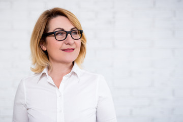 portrait of thoughtful cheerful mature business woman in eyeglasses posing over white wall