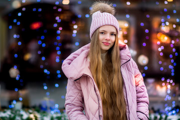 A girl in a pink warm jacket and hat with .pompon stands on the background of multi-colored blue, purple and pink garlands. Winter wonderland. Christmas and New Year concept.