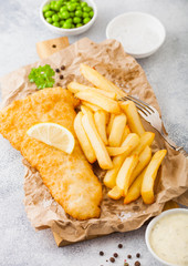 Traditional British Fish and Chips with tartar sauce on chopping board with fork and green peas on white stone table background.