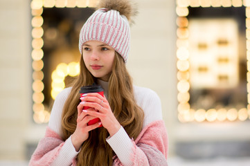 A girl in a pink warm jacket and hat with .pompon stands with coffee in the hand on the background of golden yellow lights and blurred shop windows. Winter wonderland. Christmas and New Year concept.