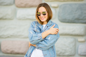 Portrait of a beautiful girl in casual white t-shirt, jeans jacket and sunglasses on the brick wall background .folded her arms