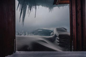 Beautiful moody winter view through a window in a mountain hut or a cottage. Winter mountain landscape seen thru window. Frozen ice, icecles, wooden house and dark snow clouds in the sky.