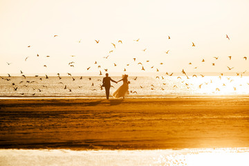 Silhouettes of the bride and groom, holding hands, run on the beach at sunset on the background of a flying flock of birds.