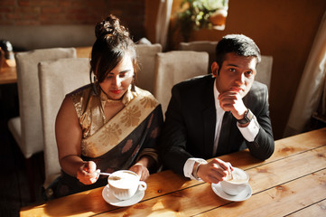 Elegant and fashionable indian friends couple of woman in saree and man in suit sitting on cafe and drinking cappuccino.