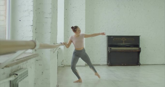 Elegant classic ballet dancer in sporty clothes rehearsing in ballet class. Beautiful ballerina performing choreographic exercises, doing rond de jamb par terre at ballet barre in dance studio.