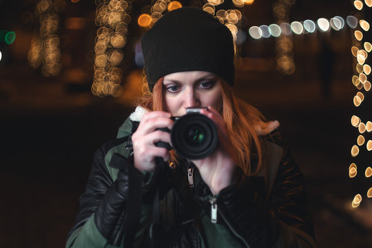 Portrait of young girl with her photo camera taking pictures in a night city street, Christmas lights on dark background