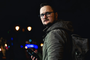 Obraz na płótnie Canvas Portrait handsome young man with a beard and mustache hold in his hand smartphone. glasses. cold season, military style clothing. on the street in the city at night