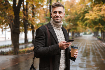 Photo of attractive man 30s wearing warm clothes walking outdoor through autumn park, and using mobile phone
