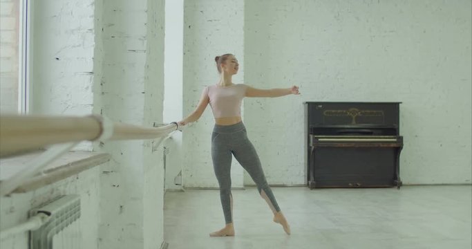 Elegant classic ballet dancer in sporty clothes rehearsing in ballet class. Beautiful ballerina performing choreographic exercises, doing rond de jamb en l'air at ballet barre in dance studio.